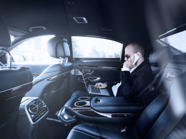 Confident young businessman working on his laptop and talking on the phone while sitting on the back seat of luxury car